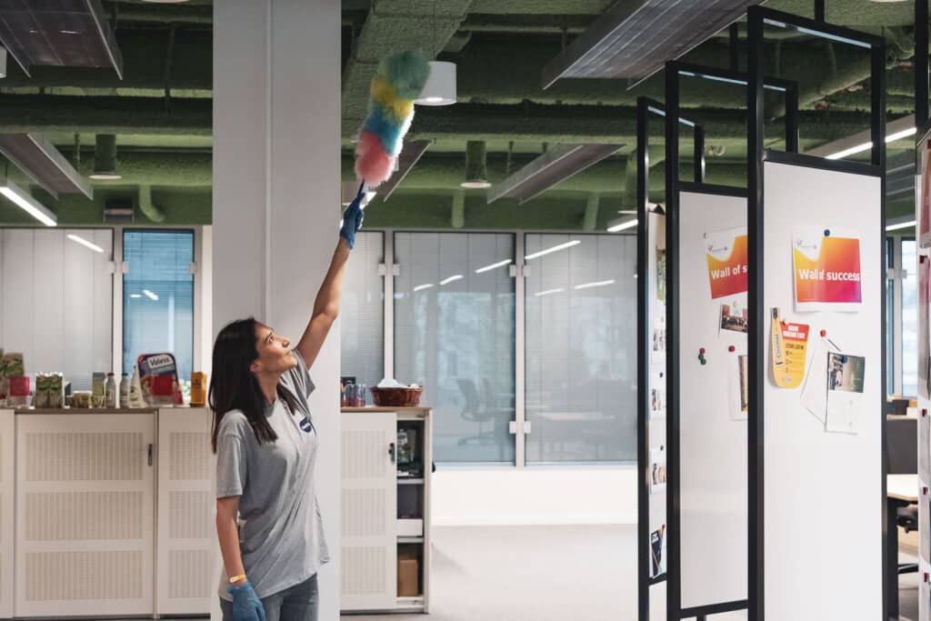 Une femme de ménage du groupe Netto nettoie les bureaux à l'aide de plumeaux colorés dans un espace de travail moderne et ouvert, décoré d'un tableau « Wall of Success ».